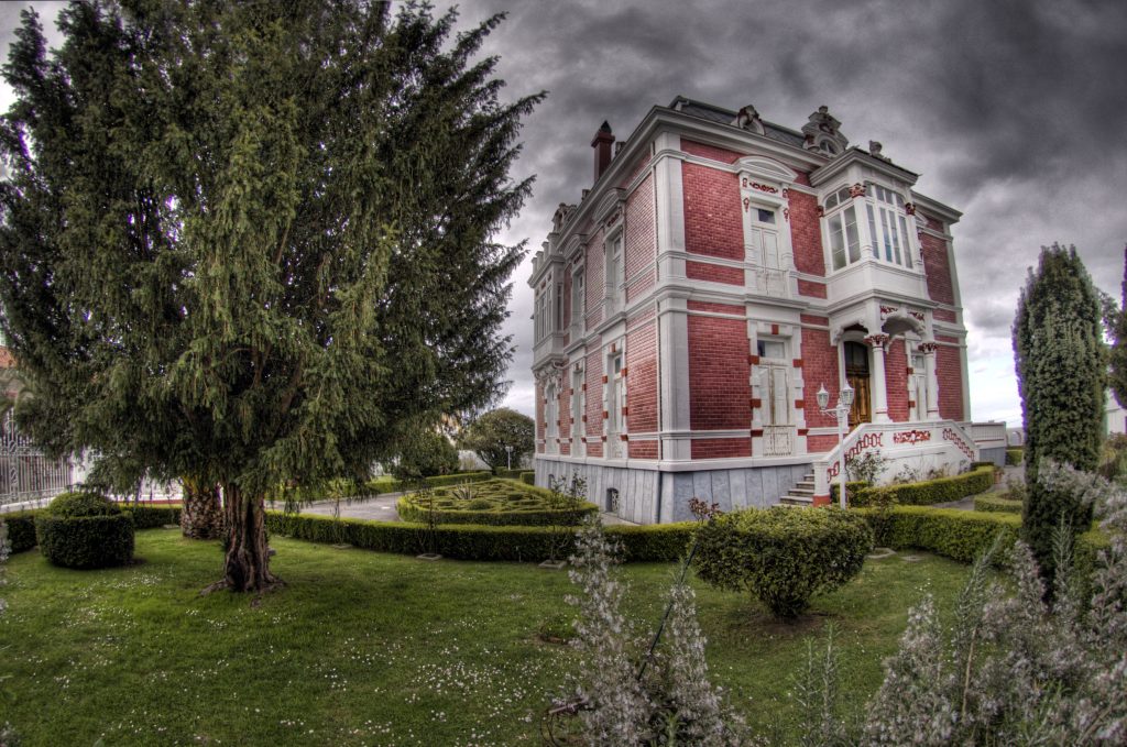 Casa indiana de Somao con fachada rojiza, galería blanca y jardín de setos